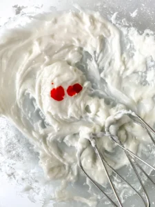 White icing mixture being prepared for homemade sprinkles with red food coloring drops on top