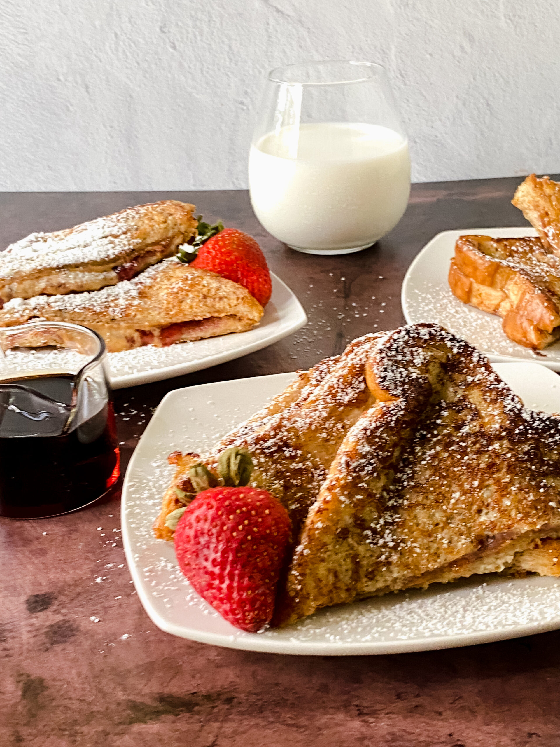 Three plates of PBJ French toast cut into triangles, each garnished with a fresh strawberry, with a glass of milk in the background. A cozy Southern-style brunch scene.