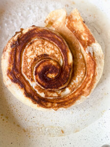 Flipped cinnamon roll pancake cooking in a skillet until the center is fully cooked.