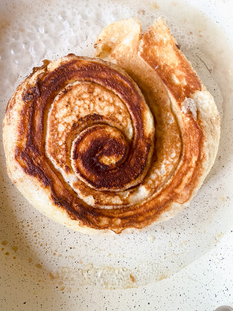 Flipped cinnamon roll pancake cooking in a skillet until the center is fully cooked.