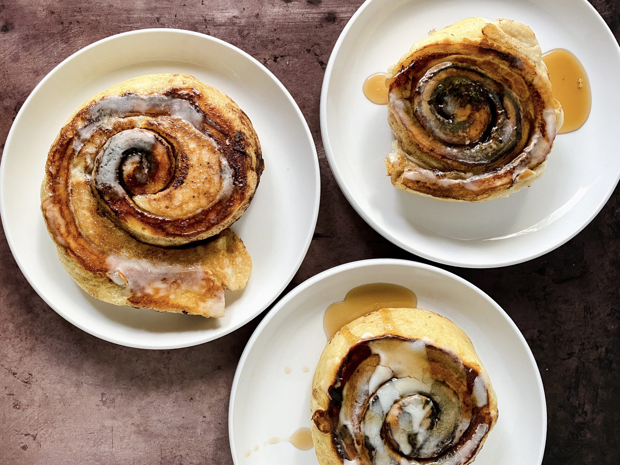 Three easy cinnamon roll pancakes topped with icing and maple syrup on white plates.