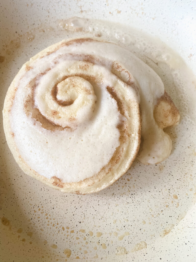 Cinnamon roll pancake cooking in a skillet with bubbles forming before flipping.