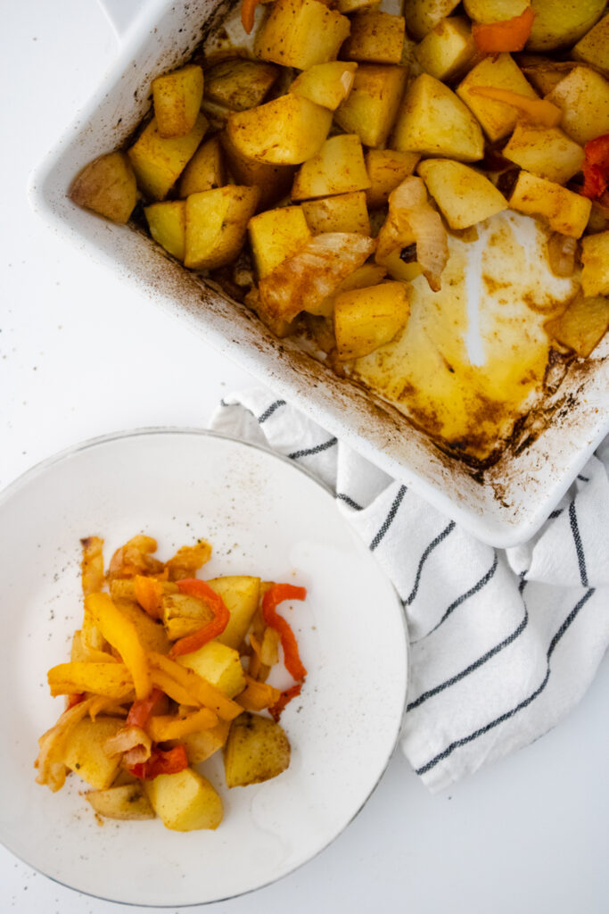 Seasoned breakfast potatoes baked with onions and bell peppers in a casserole dish with a serving plated beside it.