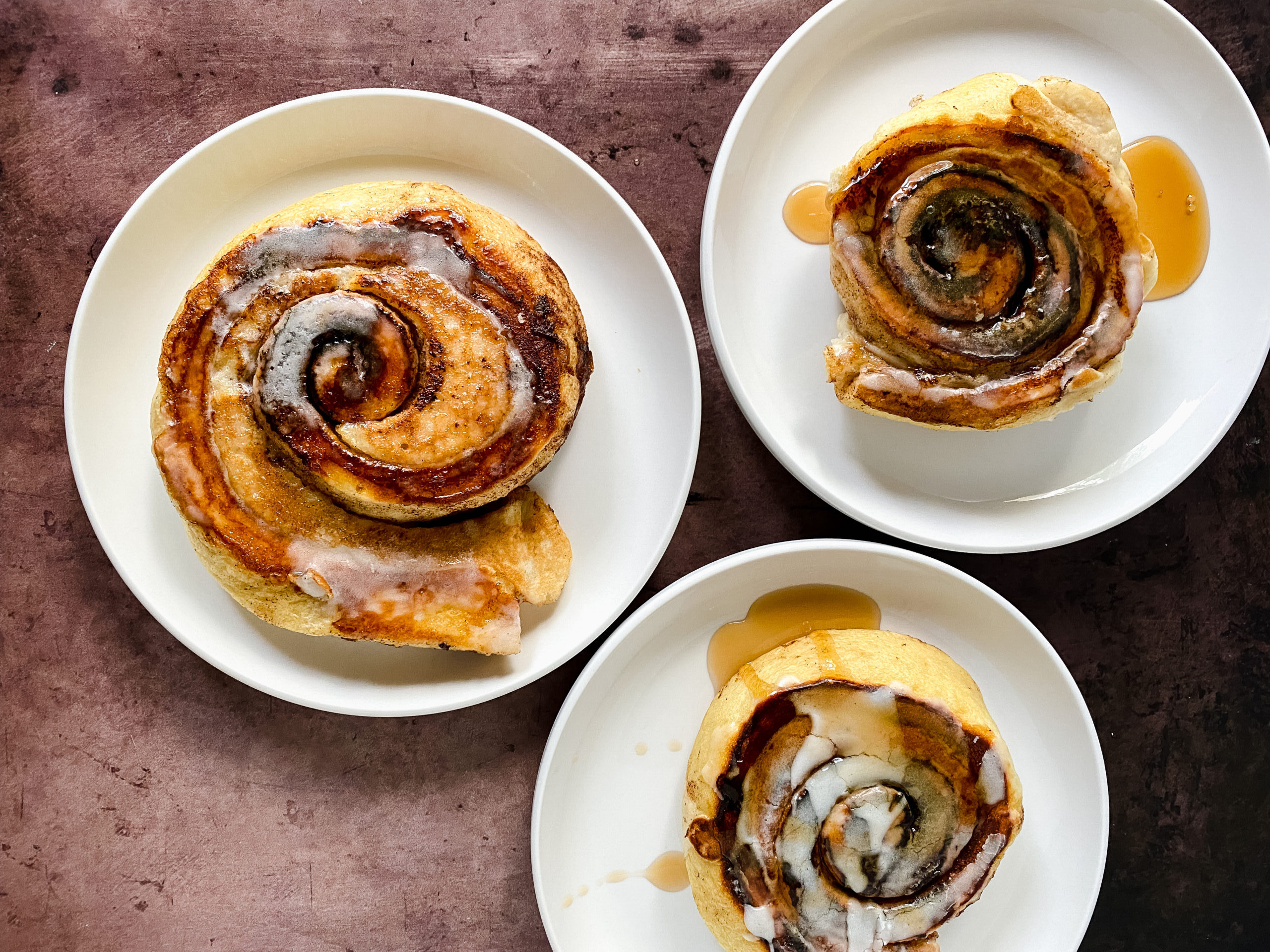 Cinnamon roll pancakes with icing drizzle served on white plates with syrup on a dark tabletop.
