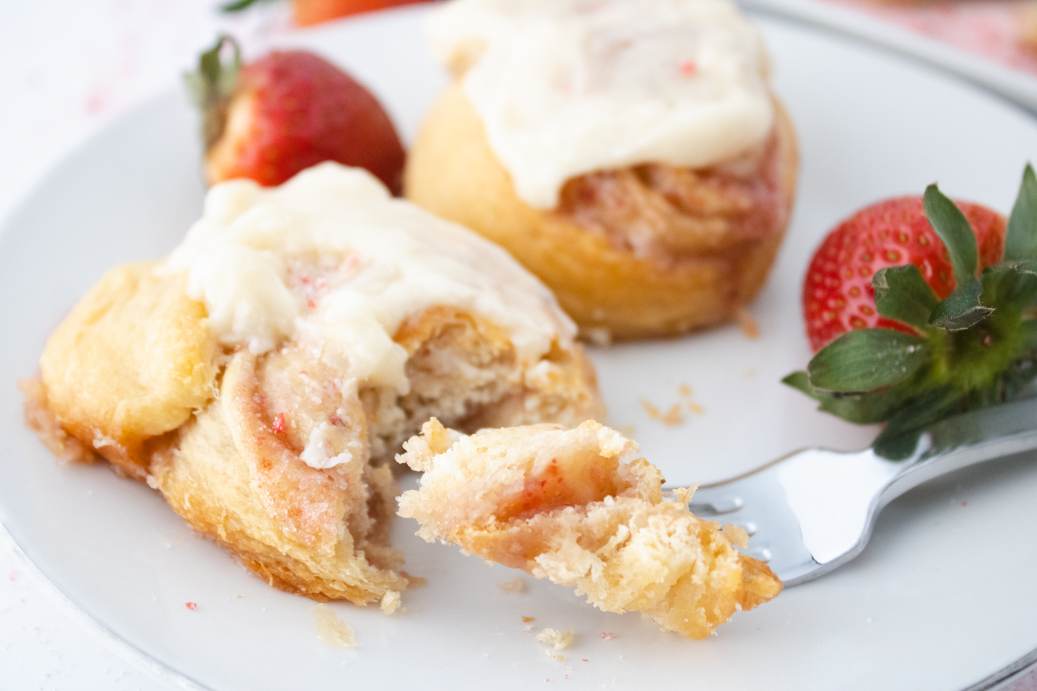 Close-up of homemade strawberry cinnamon rolls topped with cream cheese frosting and served with fresh strawberries on a white plate.