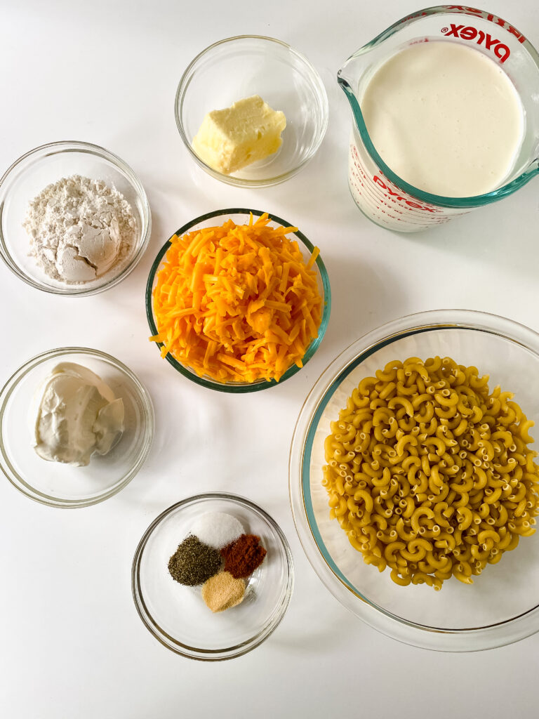 Overhead view of the ingredients for baked macaroni and cheese, including elbow macaroni, shredded cheddar cheese, milk, butter, sour cream, flour, and seasonings arranged in glass bowls on a white background.