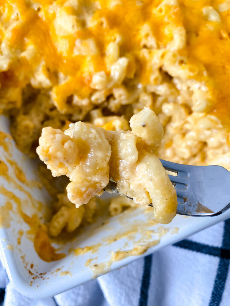 Close-up of a fork holding a bite of creamy baked macaroni and cheese with a cheesy casserole dish in the background. The image highlights the rich, smooth cheese sauce and tender pasta.