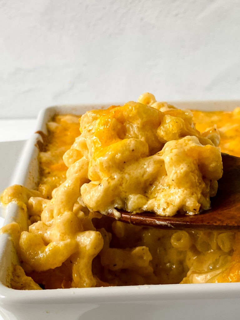 Close-up of a wooden spoon lifting a serving of creamy baked macaroni and cheese from a white baking dish. The homemade mac and cheese has a rich, cheesy sauce with a golden baked top.