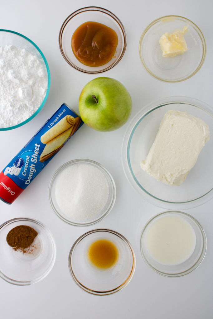 Ingredients for caramel apple Danish arranged on a white surface, including crescent sheet dough, cream cheese, apple, powdered sugar, caramel sauce, milk, butter, vanilla, sugar, and apple pie spice.