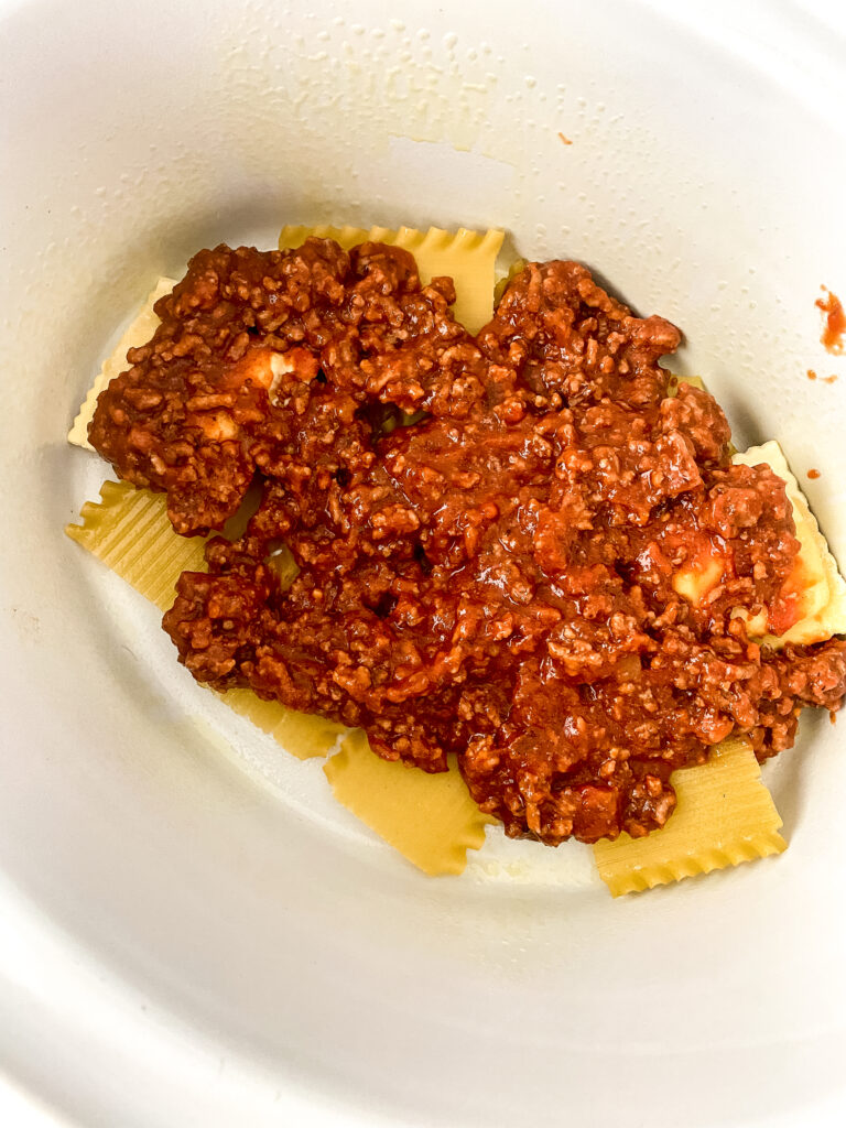 Meat sauce spooned over oven-ready lasagna noodles and frozen cheese ravioli in the first layer of easy crock pot lasagna.