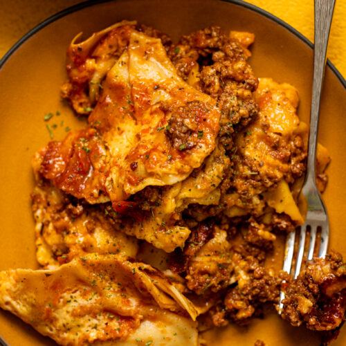 A serving of crock pot lasagna layered with ravioli, meat sauce, and melted cheese, served with crusty bread.