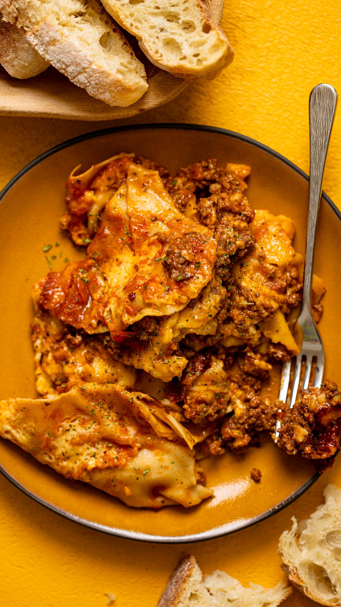 A serving of crock pot lasagna layered with ravioli, meat sauce, and melted cheese, served with crusty bread.
