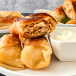 Crispy golden buffalo chicken egg rolls stacked on a plate with a bowl of ranch dressing for dipping.