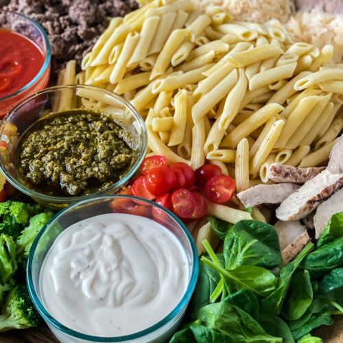 Close-up of a pasta charcuterie board featuring penne pasta, marinara sauce, Alfredo sauce, pesto, ground beef, grilled chicken, spinach, broccoli, cherry tomatoes, and shredded cheese.
