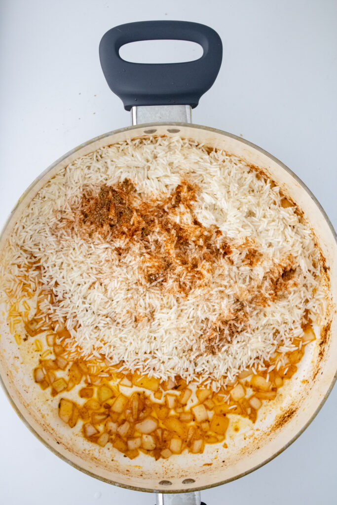 Rinsed basmati rice and reserved seasonings being added to a skillet with cooked onions for one pot chicken and rice.