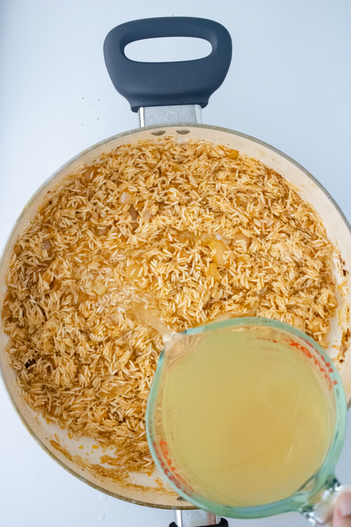 Chicken stock being poured into a skillet with seasoned basmati rice and onions for one pot chicken and rice.