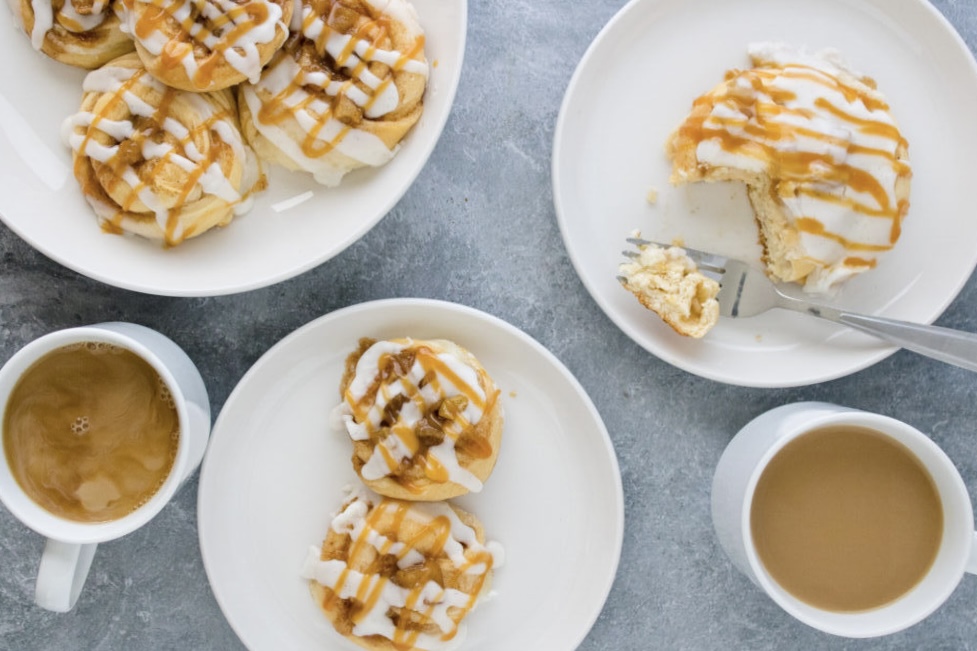 Caramel apple Danish pastries drizzled with icing and caramel sauce served with coffee on white plates.