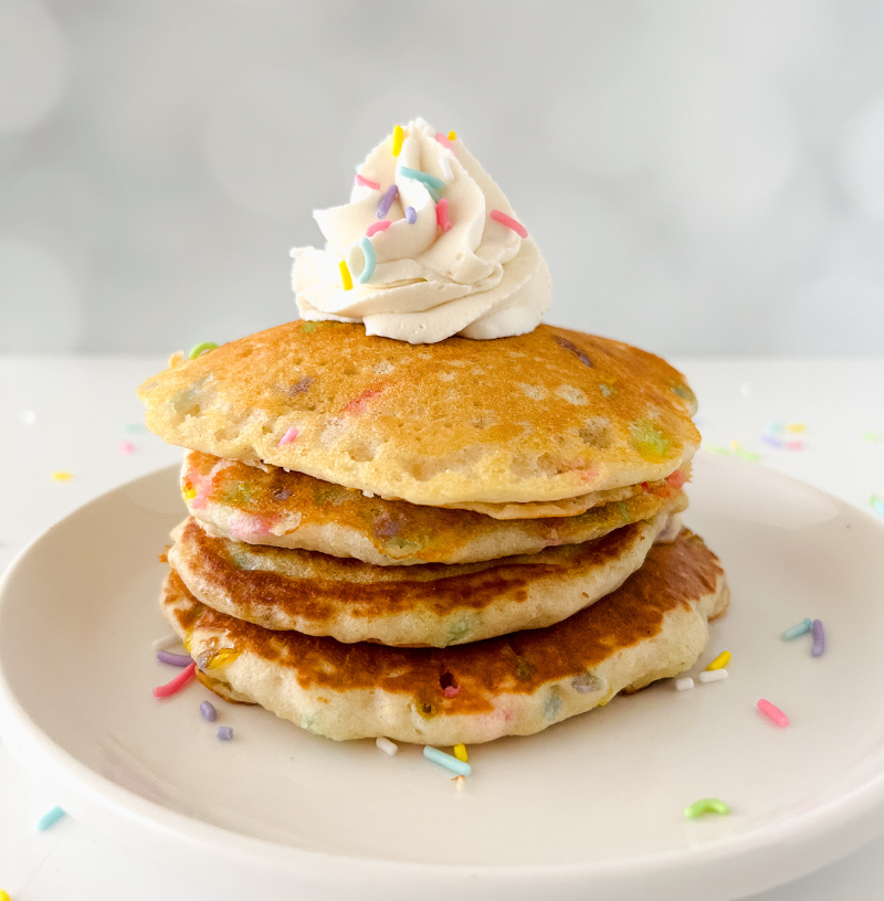 Stack of fluffy buttermilk confetti pancakes topped with whipped cream and rainbow sprinkles on a white plate.