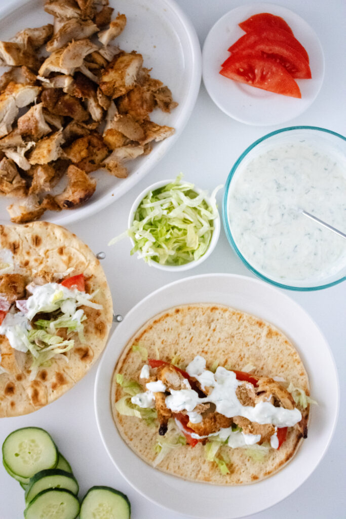 homemade chicken shawarma on pitas on a white board. photographed with sliced cucumber, lettuce, tomato and extra chicken.