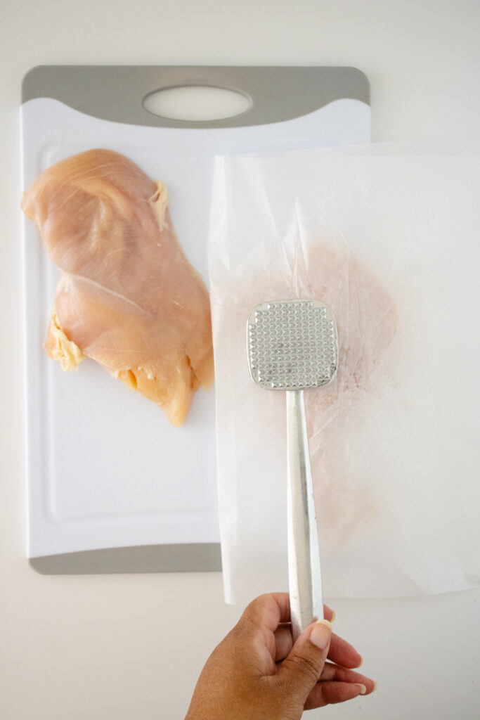 chicken cutlet on cutting board. one piece under parchment being hit with meat mallet