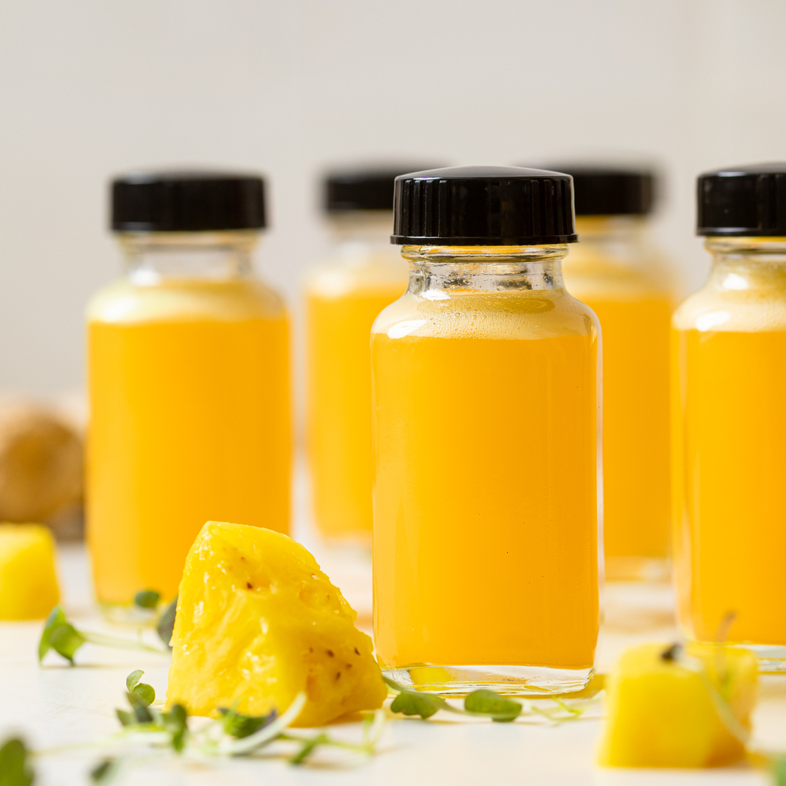 Forward-facing image of small glass bottles filled with golden pineapple ginger shots arranged in a triangle on a white surface, showcasing their bright, refreshing color.