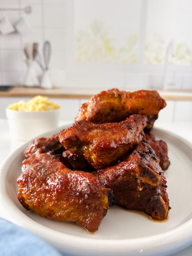 Stack of juicy air fryer baby back ribs coated in barbecue sauce on a white plate with a side of macaroni and cheese in the background.