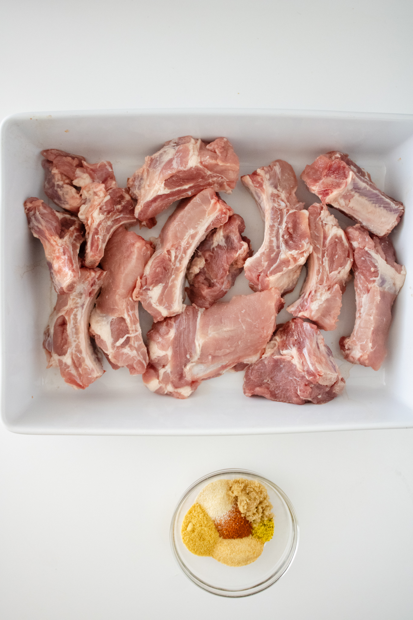 Raw baby back rib sections in a white dish with a small bowl of seasonings including BBQ seasoning, garlic powder, onion powder, ground mustard, lemon pepper, brown sugar, and pepper.
