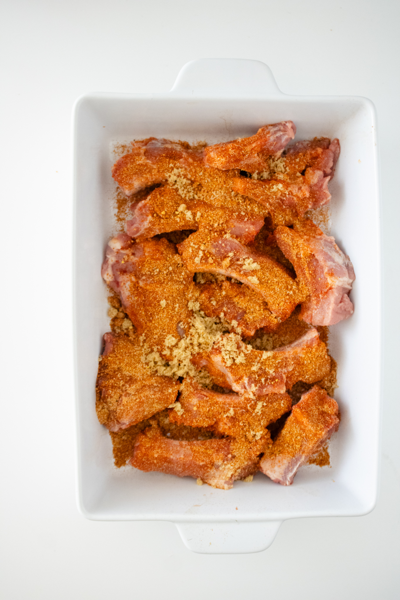 Baby back rib sections coated with BBQ seasoning, brown sugar, and spices in a white baking dish before air frying.