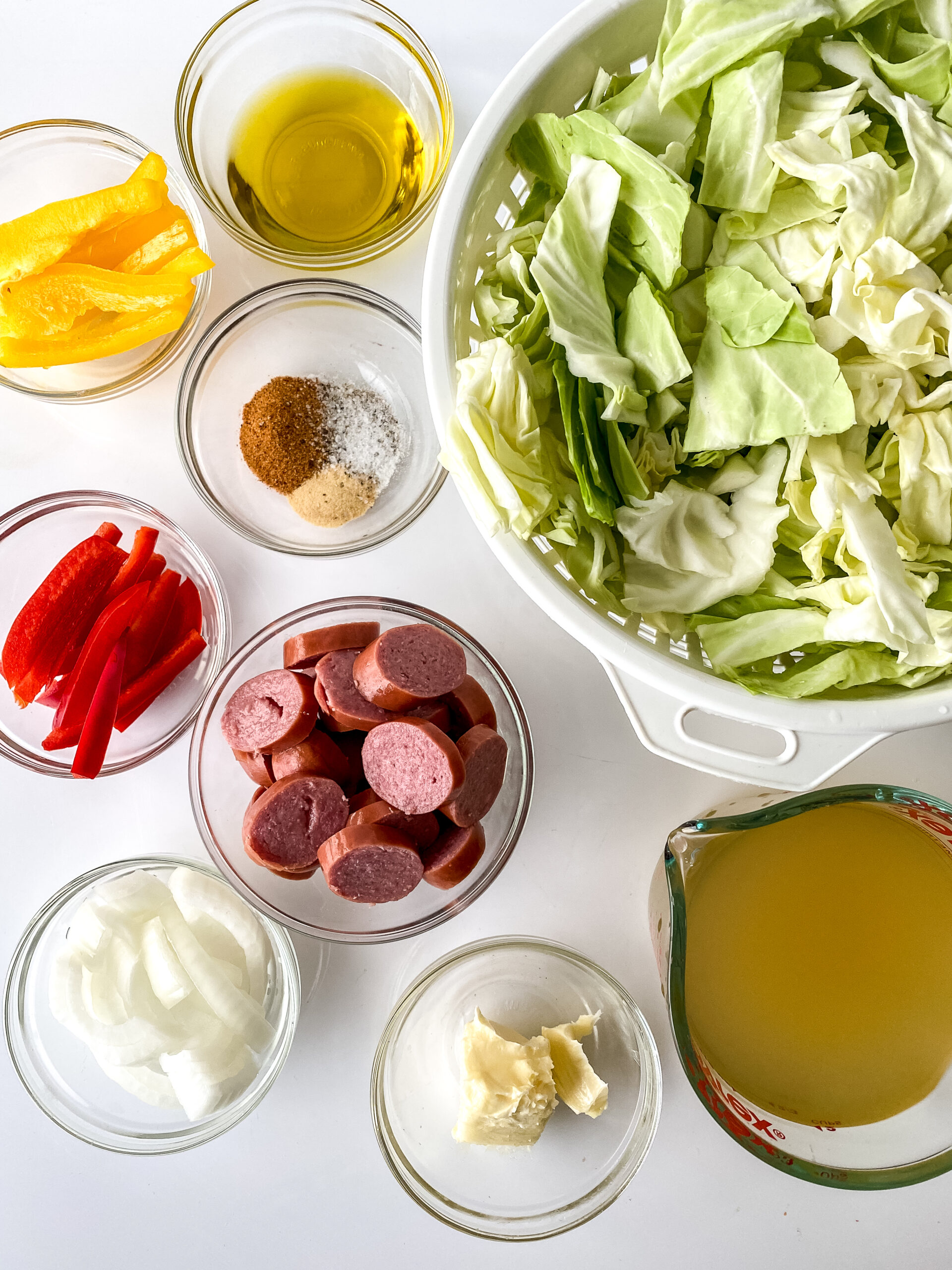 Ingredients for Southern-style cabbage and sausage arranged on a white surface, including chopped cabbage, smoked turkey sausage, bell peppers, onion, butter, oil, seasonings, and chicken stock.
