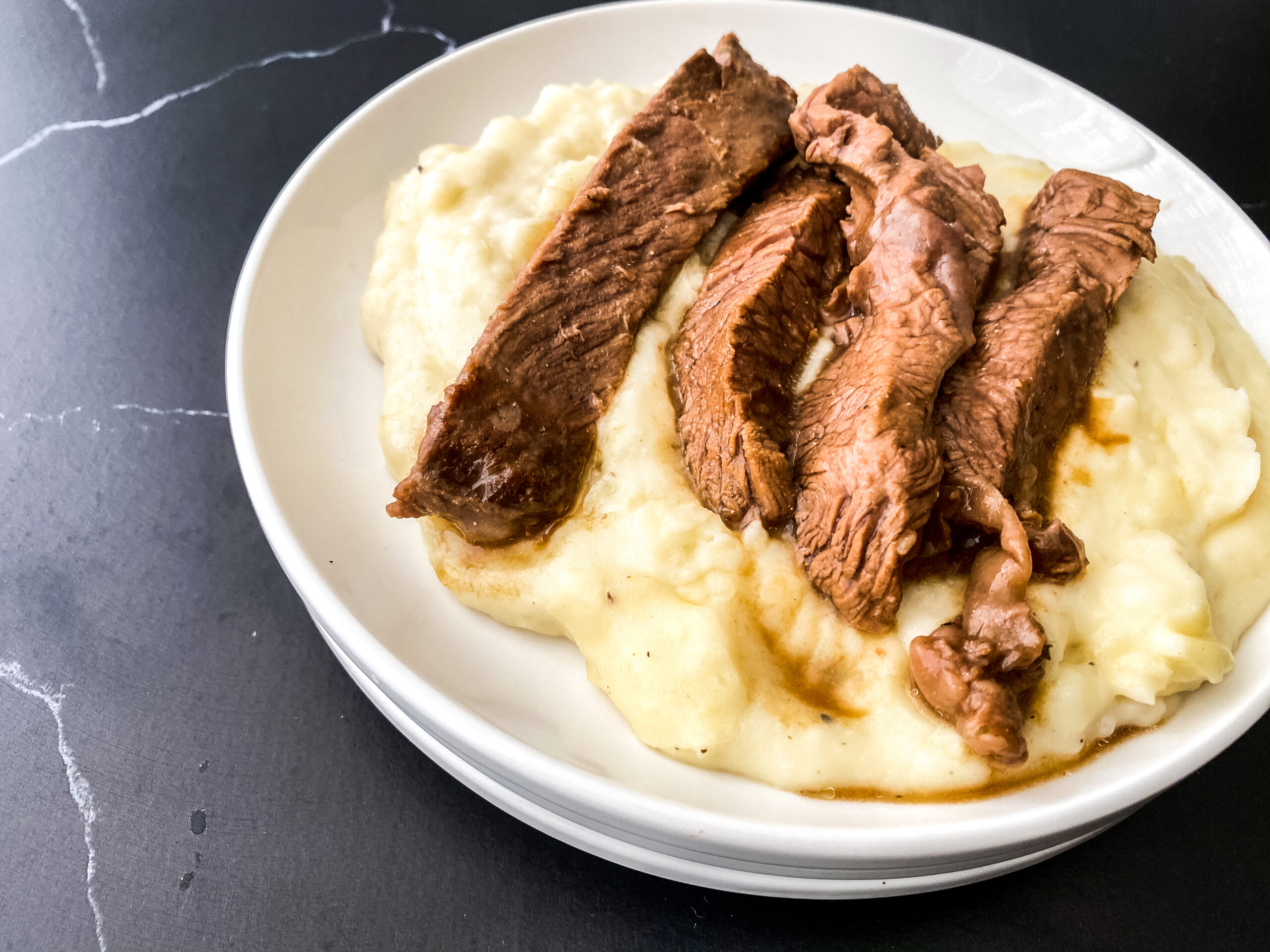 Tender crockpot sirloin steak slices served over creamy mashed potatoes with savory beef juices in a white bowl.