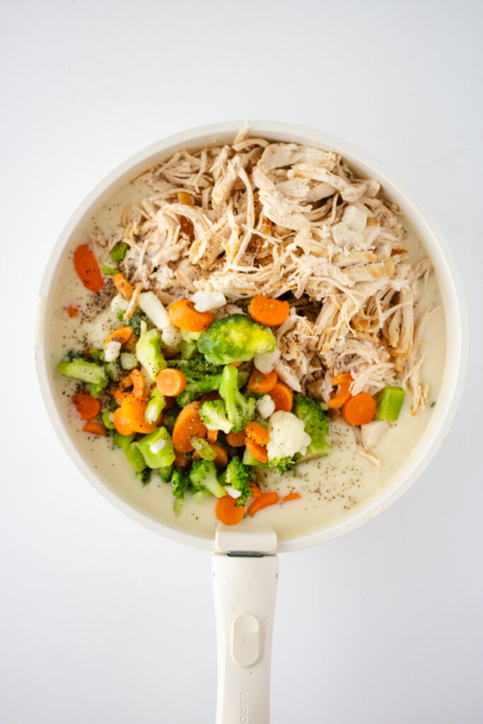 Rotisserie chicken, frozen mixed vegetables, and seasonings added to creamy chicken pot pie sauce in a skillet.