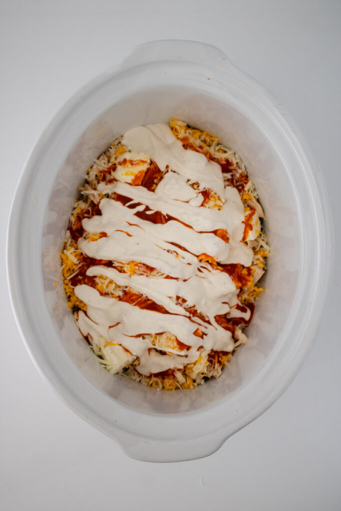 Buffalo sauce and ranch dressing poured over chicken, cream cheese, and shredded cheese in a slow cooker.