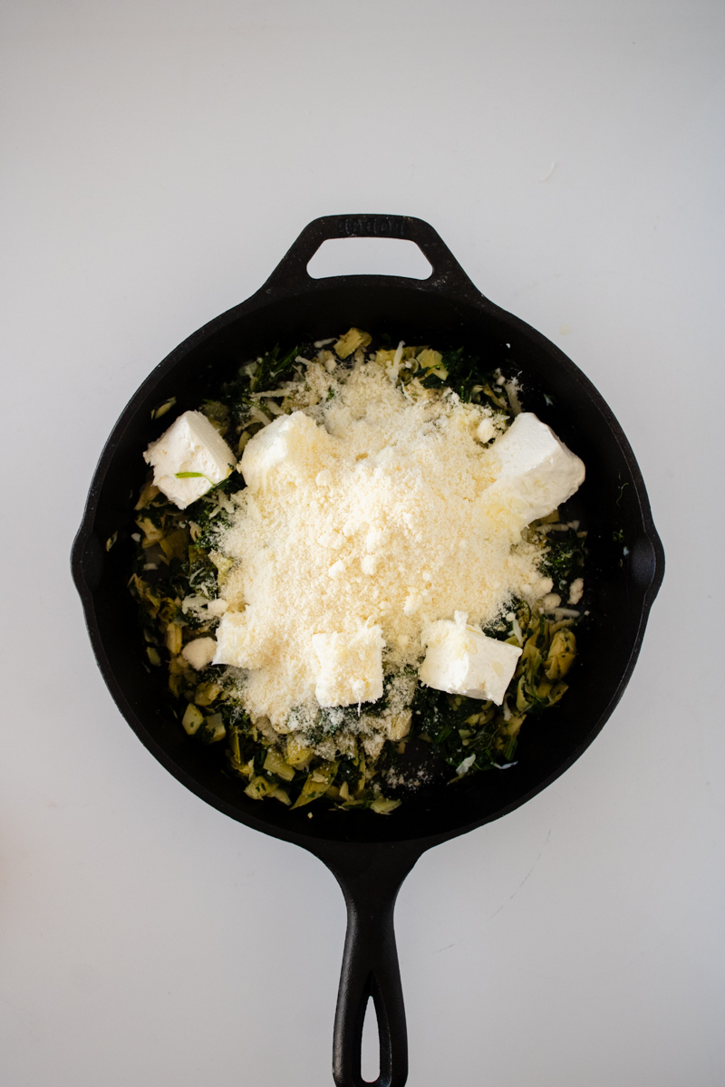 Cream cheese, mozzarella cheese, and Parmesan cheese added to spinach and artichokes in a cast iron skillet.