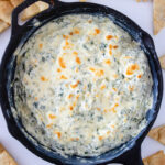 Spinach artichoke dip baked in a cast iron skillet with a golden, bubbly cheese topping and pita chips for serving.