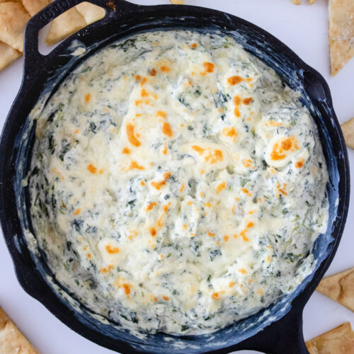 Spinach artichoke dip baked in a cast iron skillet with a golden, bubbly cheese topping and pita chips for serving.