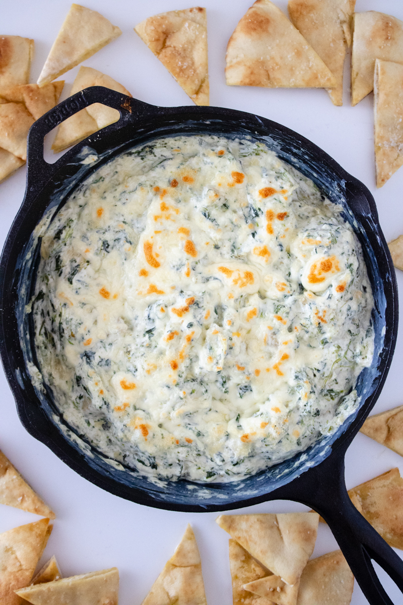 Spinach artichoke dip baked in a cast iron skillet with a golden, bubbly cheese topping and pita chips for serving.