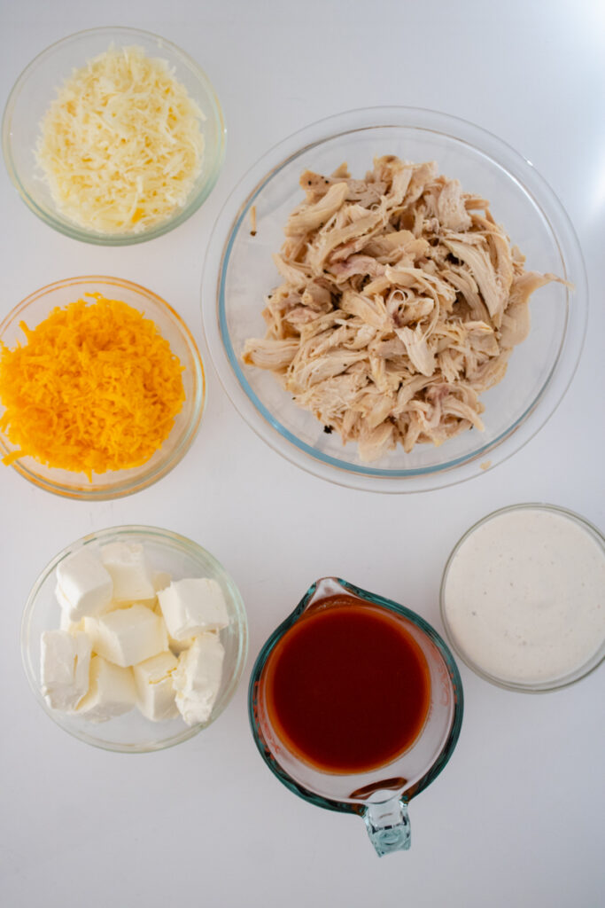 Ingredients for buffalo chicken dip including shredded rotisserie chicken, cream cheese, ranch dressing, buffalo sauce, cheddar cheese, and mozzarella cheese.