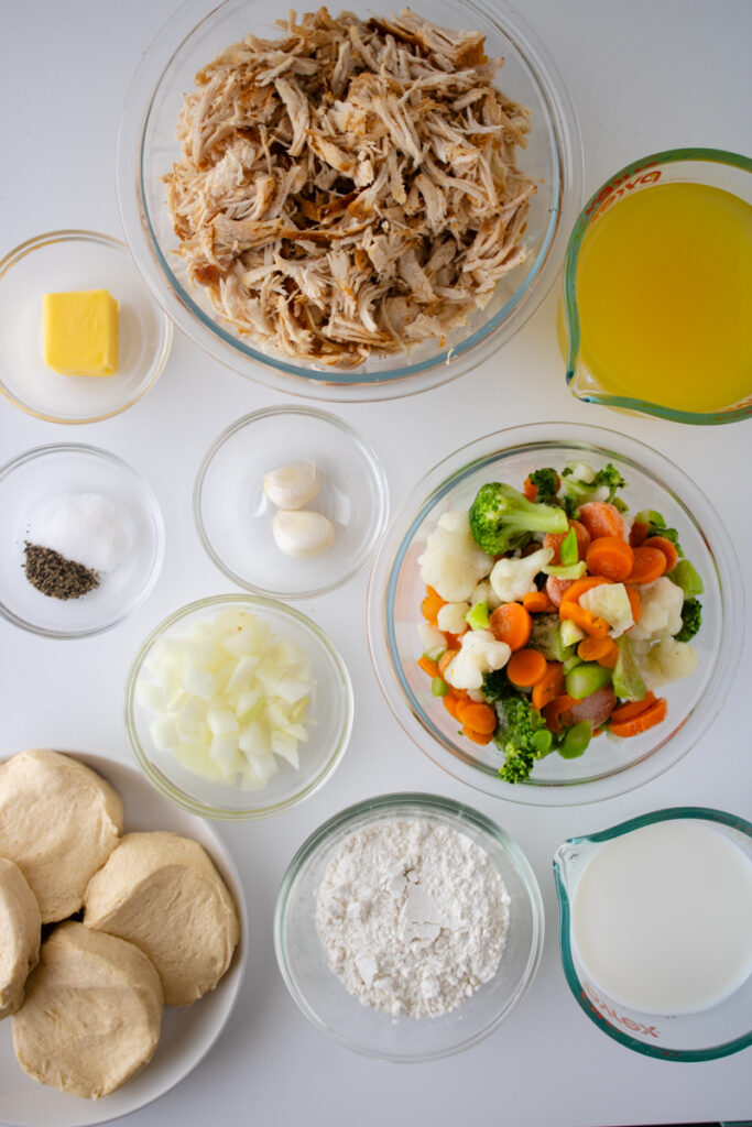 Ingredients for chicken pot pie with biscuits including rotisserie chicken, frozen mixed vegetables, chicken broth, milk, flour, onions, garlic, butter, seasonings, and refrigerated biscuit dough.