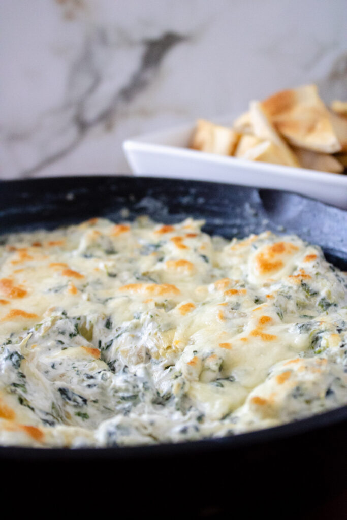 Creamy spinach artichoke dip baked in a cast iron skillet with a golden cheese topping and pita chips in the background.