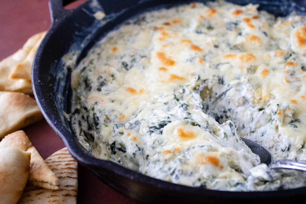 Creamy spinach artichoke dip baked in a cast iron skillet with a golden cheese topping and pita chips for serving.