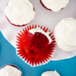 Red velvet cupcakes topped with cream cheese frosting, including one cupcake showing the moist red velvet interior.
