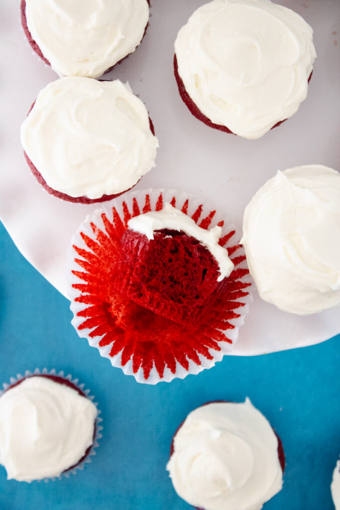 Red velvet cupcakes topped with cream cheese frosting, including one cupcake showing the moist red velvet interior.
