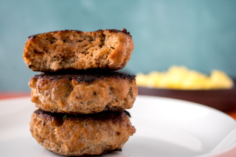 Stack of three homemade turkey sausage patties on a white plate with scrambled eggs blurred in the background.