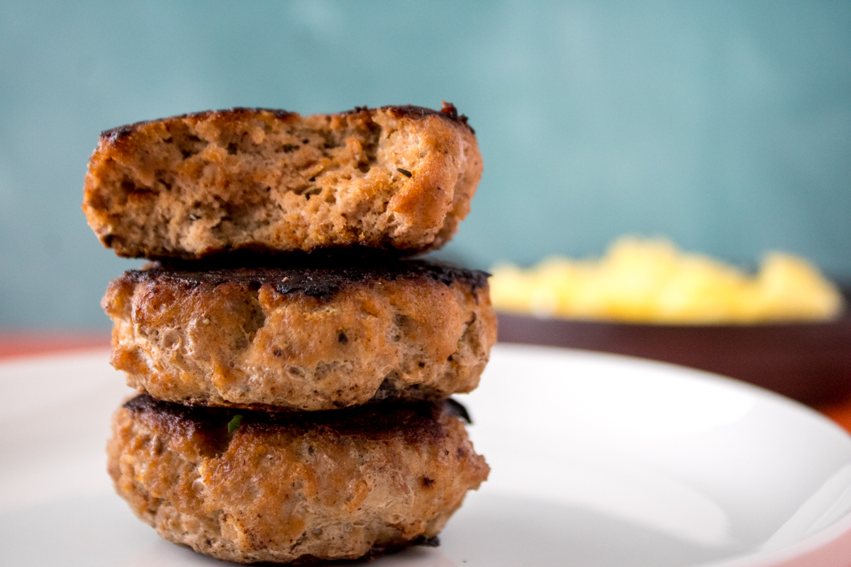 Stack of three homemade turkey sausage patties on a white plate with scrambled eggs blurred in the background.