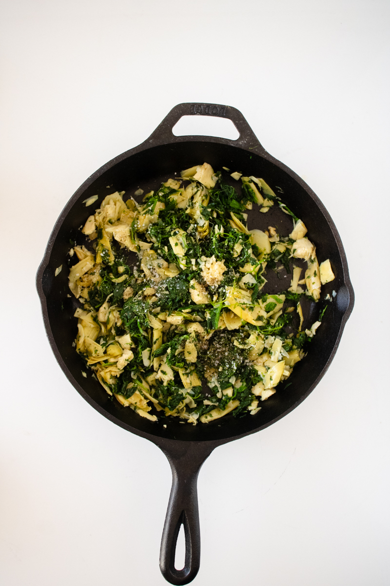 Spinach and artichokes cooking in a cast iron skillet with garlic, salt, and black pepper added.