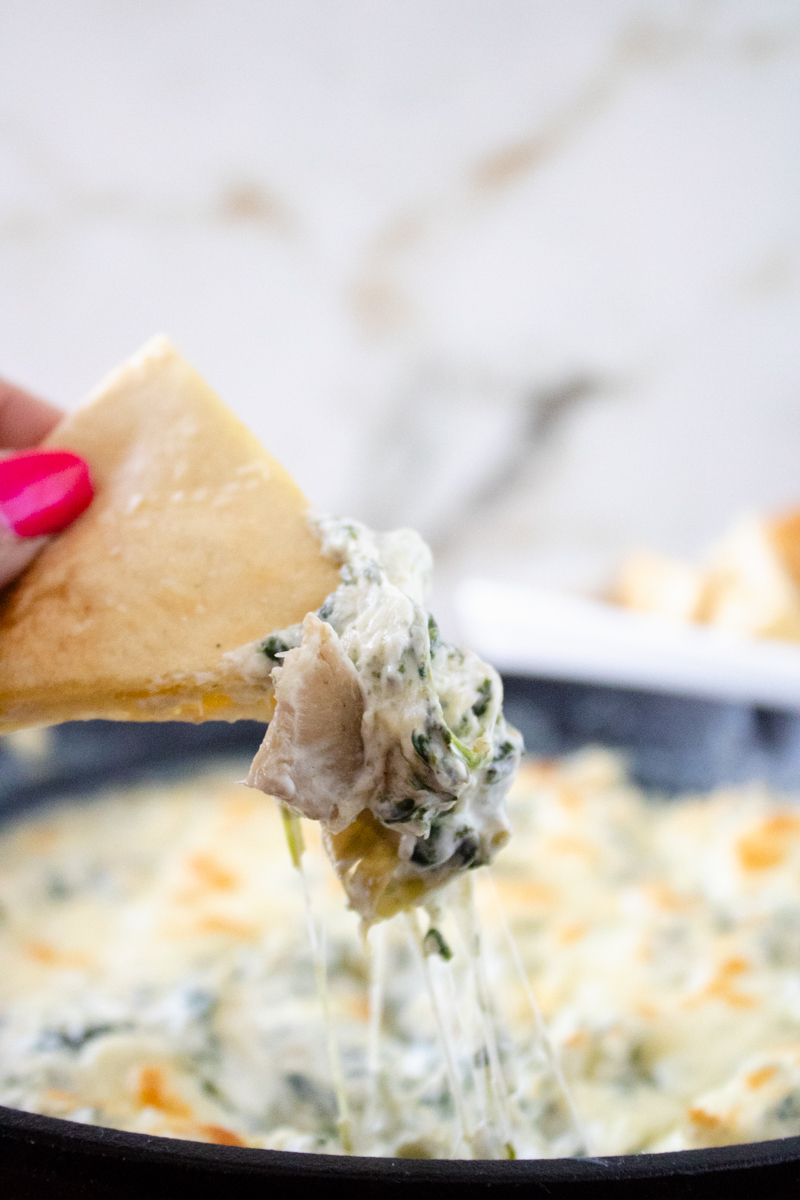 Pita chip dipped into creamy spinach artichoke dip with melted cheese stretching from a cast iron skillet.