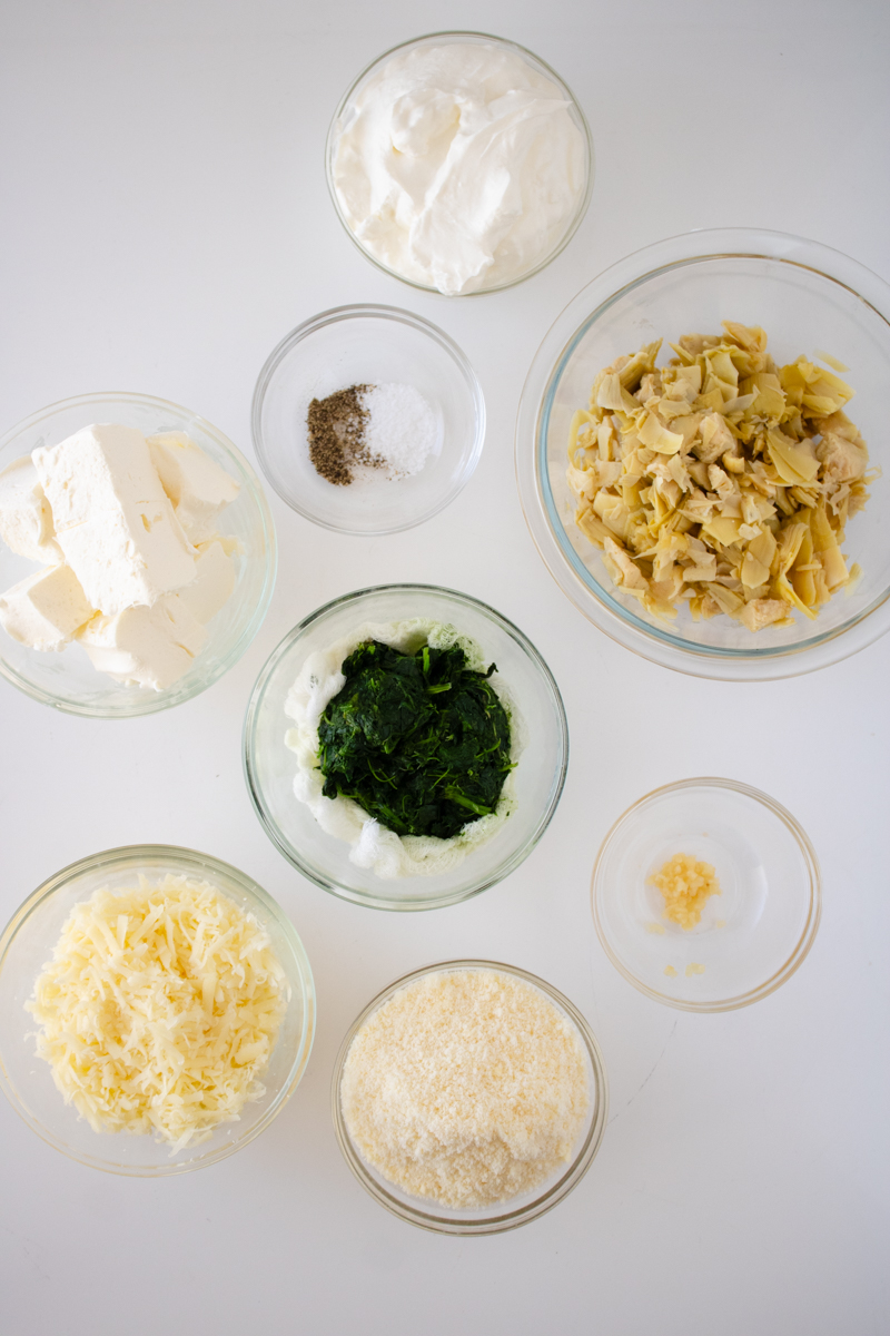 Ingredients for spinach artichoke dip including frozen spinach, artichoke hearts, cream cheese, sour cream, mozzarella, Parmesan, garlic, salt, and pepper.