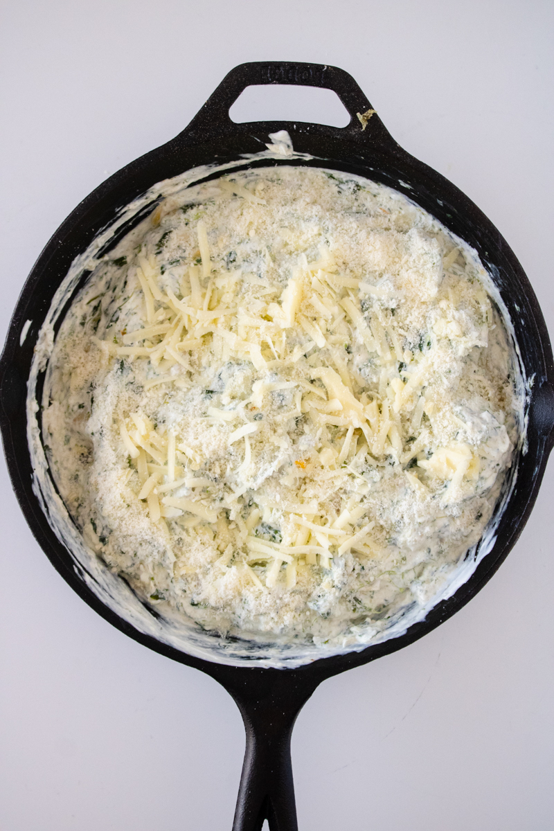 Spinach artichoke dip in a cast iron skillet topped with mozzarella and Parmesan cheese before broiling.